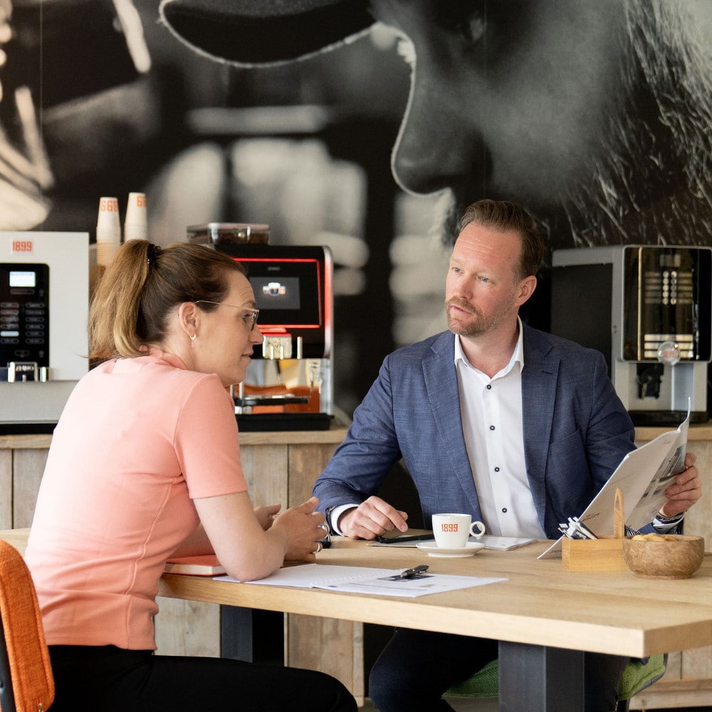 Een vrouwelijke klant kijkt geïnteresseerd naar het boekje wat de account manager haar laat zien aan tafel voor een muur met koffiemachines in de showroom van Feyen